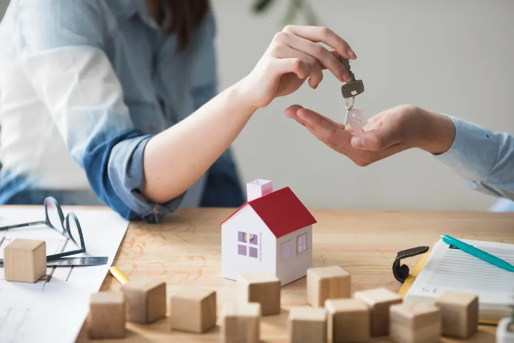One person hands another a key over a table with house model, wooden blocks, plans, and papers illustrating a real estate transaction.